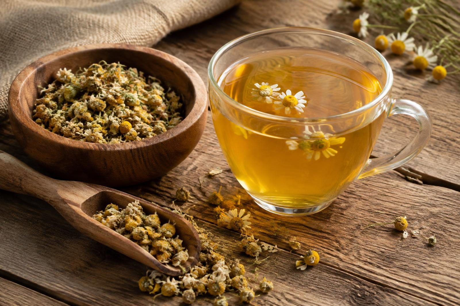 Glass cup of chamomile tea beside a wooden bowl and scoop of dried chamomile flowers on a rustic table