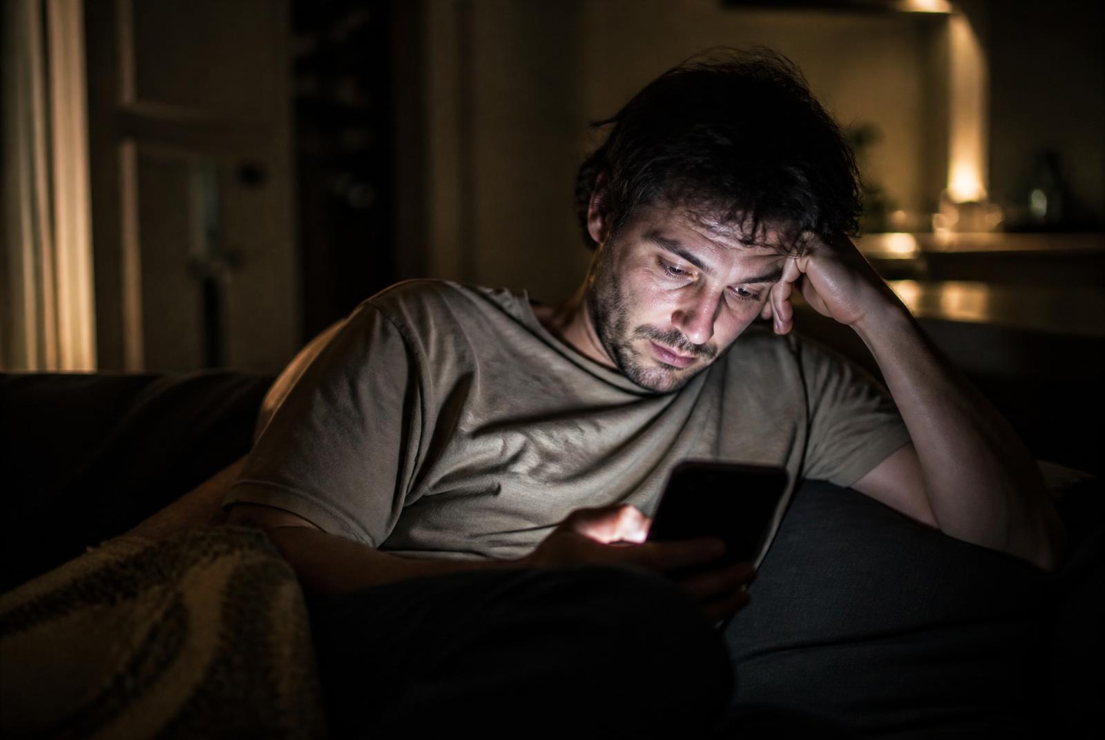 Tired man sitting alone on a couch late at night, his face lit by a phone screen, caught in an unhealthy habit loop
